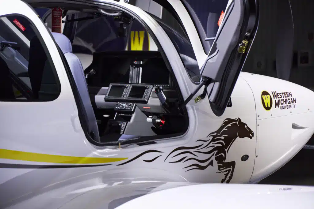 A close-up view of the cockpit door of a Western Michigan University TRAC G7 aircraft, showing its sleek white exterior with a yellow and grey stripe. The university's logo and a stylized horse design are visible on the fuselage.